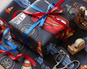 May include: A rectangular gift wrapped in dark gray paper featuring a repeating pattern of cartoon cars, orange traffic cones, and silver wrenches. The package is tied with a bright red and blue satin ribbon bow, with scissors and tape nearby.