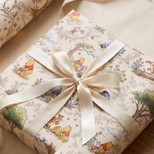 May include: A square gift box wrapped in cream-coloured paper featuring a repeating pattern of Winnie the Pooh characters, trees, and flowers. The package is tied with a smooth, cream-coloured satin ribbon tied in a bow at the centre.