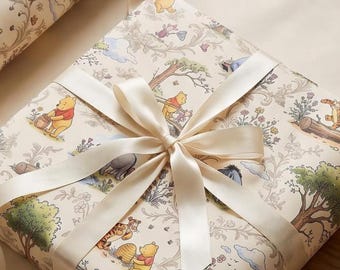 May include: A square gift box wrapped in cream-colored paper featuring a repeating pattern of Winnie the Pooh characters, trees, and flowers. The package is tied with a smooth, cream-colored satin ribbon tied in a bow at the center.