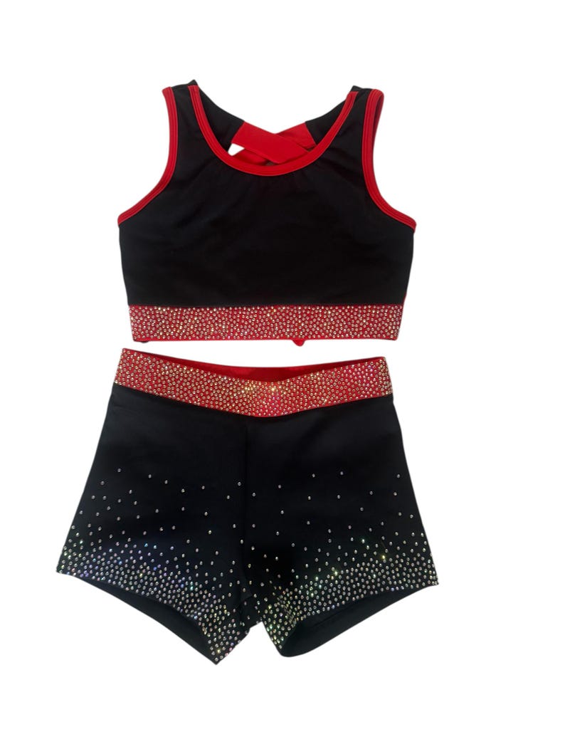 Peut inclure: Ensemble d'uniforme de cheerleading noir avec garniture rouge et embellissements scintillants. Le haut est un crop top avec un n&oelig;ud rouge dans le dos. Le short a une ceinture rouge assortie et est recouvert de strass scintillants.
