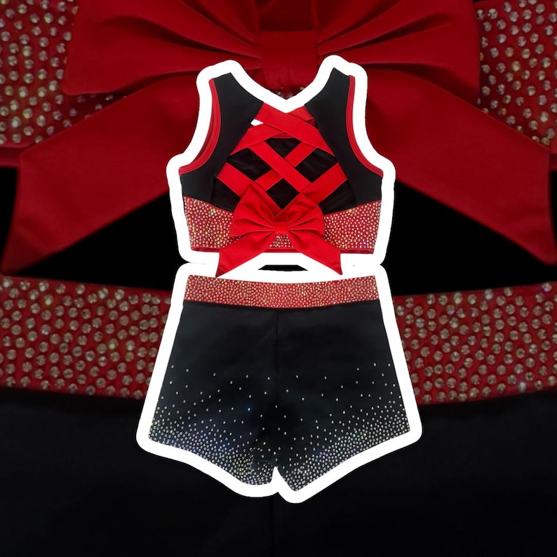 Peut inclure: Ensemble uniforme de cheerleading noir et rouge. Le haut pr&eacute;sente un dos crois&eacute; et un n&oelig;ud rouge. Le short est noir avec des strass. Le haut et le short ont une bordure rouge. Le fond est noir avec un n&oelig;ud rouge.
