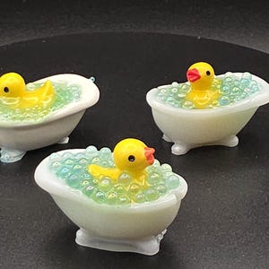 May include: Three miniature bathtubs with yellow rubber duckies inside. The tubs are white with light blue accents, filled with blue bubbles. The duckies have orange beaks and black eyes, creating a playful and whimsical scene.