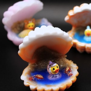 Op de afbeelding: Schelpenbeeldjes met een eend en vissen erin. De schelpen zijn wit en oranje, met een blauw zwembad erin. De eend heeft een gele snavel en een witte hoed. De vissen zijn rood en goud.