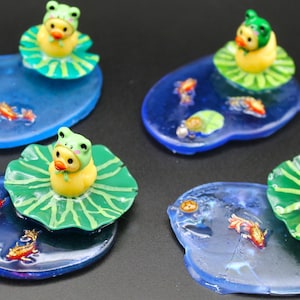 May include: Four miniature resin pond dioramas, each featuring a yellow duckling wearing a green frog hat perched on a green lily pad. The blue translucent bases contain embedded orange and white koi fish figures and small green lily pads.