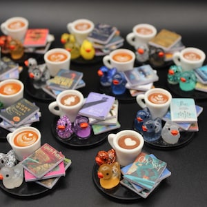 Könnte beinhalten: Miniatur-Kaffeetassen mit Latte Art, jeweils auf einem schwarzen Untersetzer, umgeben von kleinen Büchern und Entenfiguren. Die Enten sind in verschiedenen Farben, darunter Gelb, Blau und glitzerndes Lila. Eine charmante, skurrile Sammlung.