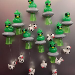 Peut inclure: Une collection d'aimants représentant des figurines d'extraterrestres verts assis sur des ovnis gris avec des rayons tracteurs verts. Chaque ovni est relié par un fil fin à une petite figurine de vache blanche et noire aux yeux noirs et becs jaunes.