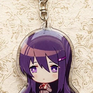 Może przedstawiać: Przezroczysty akrylowy brelok do kluczy z postacią z kresk&oacute;wki z fioletowymi włosami i oczami. Postać ma na sobie mundur szkolny. Słowo "Yuri" jest wydrukowane w lewym g&oacute;rnym rogu. Brelok ma srebrne zapięcie.