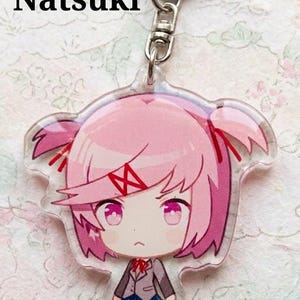 Może przedstawiać: Przezroczysty akrylowy brelok z r&oacute;żowowłosą postacią z anime o imieniu Natsuki. Postać ma r&oacute;żowe oczy, mundur szkolny i surowy wyraz twarzy. Brelok zawiera srebrny zatrzask.