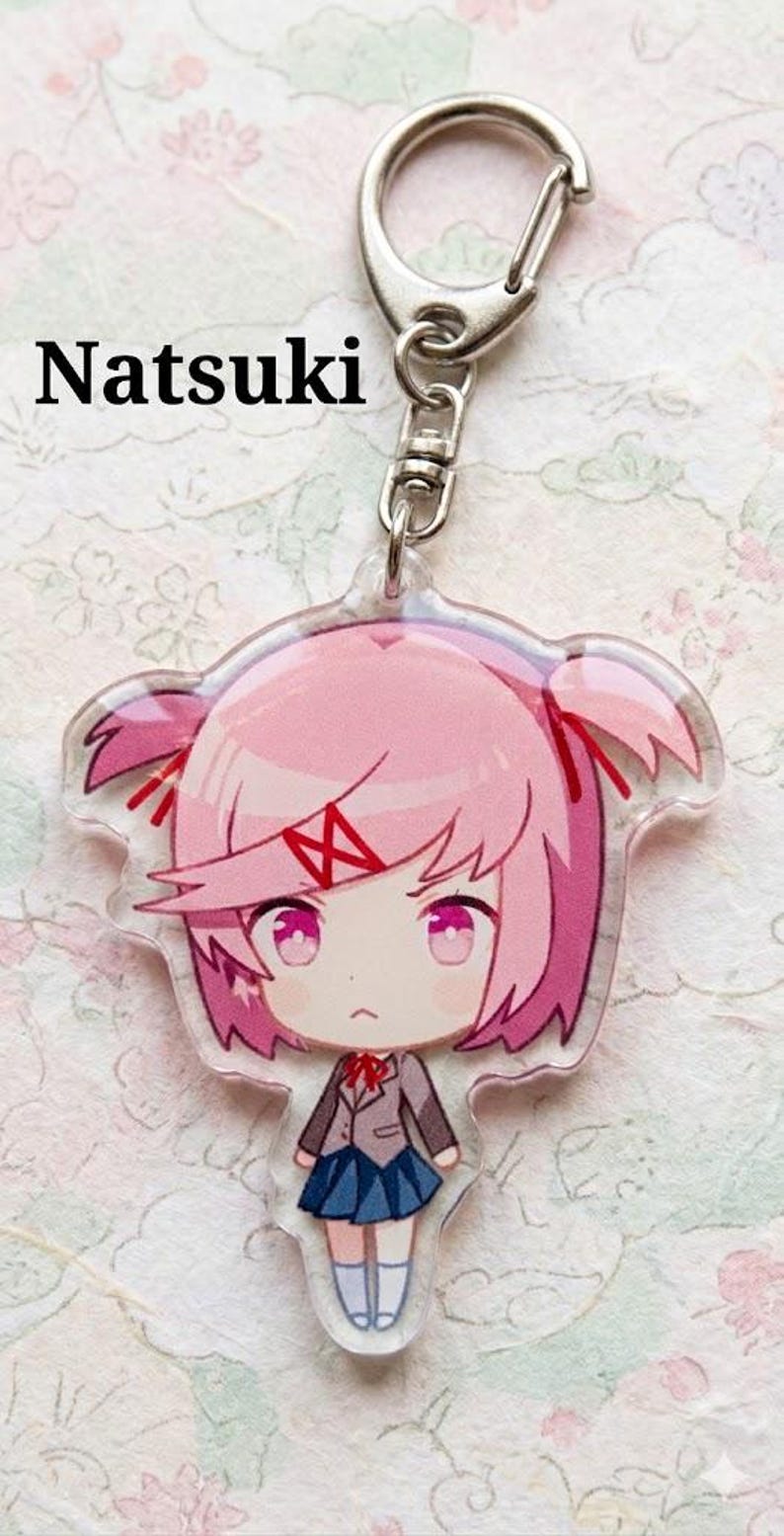 Może przedstawiać: Przezroczysty akrylowy brelok z r&oacute;żowowłosą postacią z anime o imieniu Natsuki. Postać ma r&oacute;żowe oczy, mundur szkolny i surowy wyraz twarzy. Brelok zawiera srebrny zatrzask.