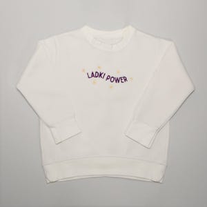 Può includere: Una felpa bianca con la scritta ricamata "LADKI POWER" in viola, con piccoli accenti floreali gialli. Il top girocollo a maniche lunghe è realizzato in un materiale morbido.