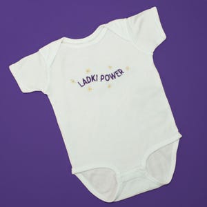 Può includere: Body da neonato bianco a maniche corte, con la scritta ricamata "LADKI POWER" in viola, ornata da piccoli fiori gialli. L'indumento è appoggiato su uno sfondo viola.