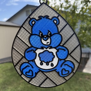 May include: A teardrop-shaped suncatcher featuring a blue bear with a white belly patch depicting a rain cloud with falling raindrops. The bear is outlined in black and set against a background of diagonal, translucent, textured gray panels.