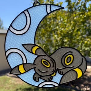 May include: A decorative hanging ornament featuring a dark gray creature with yellow ring markings resting on a light blue crescent moon. The design has a grid-like texture, black outlines, and a circular loop at the top for suspension.