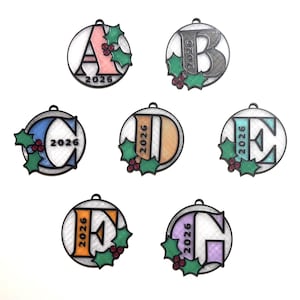 May include: A collection of seven circular ornaments featuring the letters A through G in various colors. Each ornament includes the year 2026, a black border, and green holly leaves with red berries. The letters are filled with textured patterns.