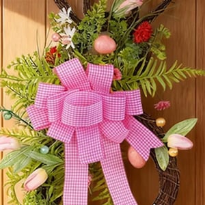May include: Easter bunny-shaped wreath made of twigs, adorned with greenery, pink tulips, and small pink eggs. A large pink and white gingham bow is centered on the wreath. The wreath hangs on a wooden door.