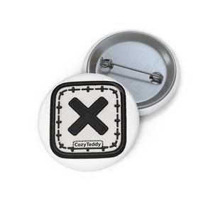 May include: A round pin-back button featuring a white square patch with a thick black border and a black X in the center. The patch design includes black stitched details around the edges and the text CozyTeddy printed in black at the bottom.
