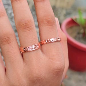 Peut inclure: Deux bagues en métal de couleur cuivre portées sur les doigts. La bague la plus large présente un motif en relief répétitif de petits cercles et de formes ovales. La bague plus fine affiche un design géométrique texturé avec des motifs.