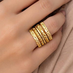 Può includere: Un set di quattro anelli in metallo color oro indossati insieme su un dito. Ogni anello presenta un motivo in rilievo distinto, inclusi disegni floreali, linee geometriche e superfici testurizzate, creando un aspetto sovrapposto e lucido.