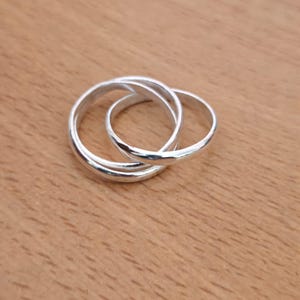 May include: A silver-coloured interlocking ring set consisting of three thin, smooth, polished bands that are looped together. The metal has a reflective surface, creating a continuous circular design where the three individual bands overlap.