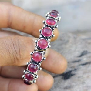 Puede incluir: Un brazalete plateado con siete piedras preciosas redondas de color rosa intenso. El brazalete tiene un diseño detallado con pequeños detalles en relieve entre cada piedra preciosa, creando una estética vintage o antigua.