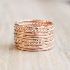 May include: A stack of seven rose gold rings featuring alternating textures. The set includes smooth bands, hammered bands with a dimpled surface, and twisted rope-style bands, all stacked vertically to create a single cohesive unit.