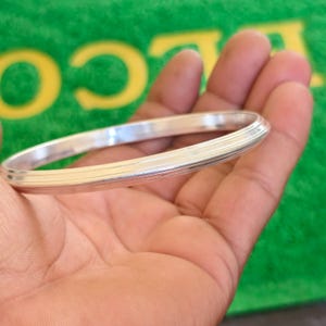 Könnte beinhalten: Ein silberfarbenes Armreif-Armband mit gerilltem Design. Das Armband wird in einer Hand gehalten, wodurch seine polierte Oberfläche und sein schlichter, eleganter Stil hervorgehoben werden. Ein klassisches Schmuckstück.