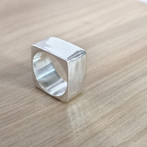 May include: A silver-colored, square ring with a brushed metal finish. The ring has a solid, rectangular top and a thick band. The ring is displayed on a light brown wooden surface.
