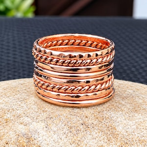 Puede incluir: Una pila de siete anillos de oro rosa con texturas alternas, que incluyen bandas lisas, diseños de cuerda trenzada y acabados martillados. Los anillos son metálicos y reflectantes, mostrando un tono cálido de color cobre en todo el conjunto.