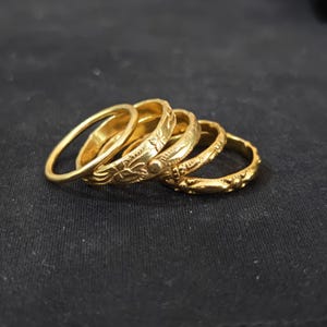 Può includere: Un set di cinque anelli in metallo color oro disposti in fila. Gli anelli presentano varie texture e motivi, tra cui fasce lisce, incisioni floreali e piccoli accenti circolari sulle superfici esterne delle fasce metalliche.