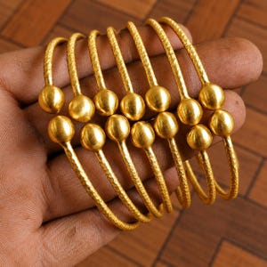 Op de afbeelding: Een set van zes goudkleurige metalen armbanden, elk met een getextureerd, gehamerd oppervlak en twee gladde, bolvormige uiteinden. De armbanden zijn open, flexibel en in een stapel gerangschikt om de metalen afwerking goed te tonen.