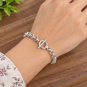 Op de afbeelding: Een zilverkleurige kettingarmband met een reeks ronde, in elkaar grijpende schakels en een opvallende knevelsluiting. De armband is gemaakt van gepolijst metaal en toont een consistent patroon van cirkelvormige schakels met een T-balk.