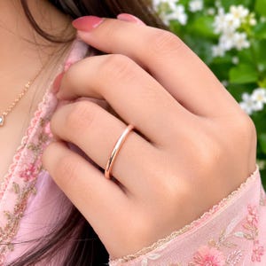 Può includere: Un semplice anello a fascia in oro rosa lucido con una superficie liscia e arrotondata. L'anello presenta un design minimalista con una finitura metallica molto brillante, indossato su un dito. La fascia ha una larghezza sottile e costante.