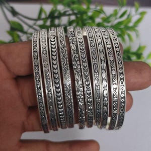 May include: A set of nine silver-toned metal bangles, each featuring a distinct embossed pattern including hearts, floral motifs, geometric lines, and paisley shapes. The bracelets have a polished finish and are arranged side-by-side in a row.