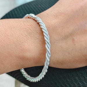 Puede incluir: Una pulsera rígida de tono plateado pulido que presenta un diseño de cuerda trenzada continua. La superficie metálica es reflectante y lisa, formando una figura circular que rodea la muñeca con una textura en espiral uniforme en toda la banda.