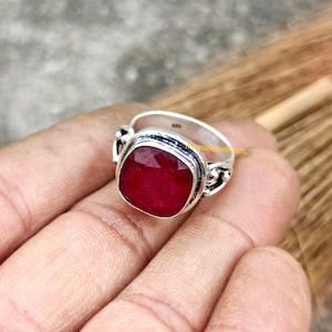Op de afbeelding: Een zilveren ring met een grote, vierkant geslepen, dieprode edelsteen. De edelsteen is gezet in een bezel setting en de ring heeft decoratieve filigraan details aan de zijkanten. De ring wordt op een hand getoond.