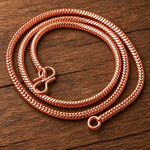 Op de afbeelding: Een koperkleurige slangenketting met een glad, flexibel en afgerond schakelontwerp. De ketting is voorzien van een opvallend haak-en-oogsluitingsmechanisme aan de uiteinden en heeft een metallic afwerking met een warme roodbruine tint.