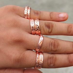 May include: A collection of seven copper-toned metal rings worn on the fingers. Each ring features a distinct embossed pattern, including floral motifs, geometric shapes, and linear textures. The bands have a polished finish and varying widths.