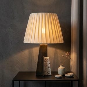 May include: A cream-colored pleated lampshade sits atop a dark bronze-colored lamp base. The lamp is on a black table, with a small vase and decorative stones. The lamp's design is modern and elegant.