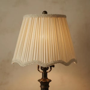 Könnte beinhalten: Eine cremefarbene Lampe mit einem plissierten Lampenschirm. Der Lampenschirm hat einen gewellten Rand mit Zierleiste. Der Lampenfuß ist dunkelbraun mit Bronze-Finish. Die Lampe ist eingeschaltet und das Licht scheint durch den Lampenschirm.