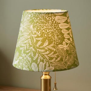 May include: A conical lamp shade featuring a vibrant green background with a detailed white botanical print of various leaves and ferns. The shade is mounted on a polished brass base with a matching brass pull chain for operation.