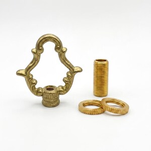 May include: A brass lamp finial featuring an ornate, decorative loop design with a threaded base, accompanied by a separate threaded metal pipe nipple and two matching circular knurled nuts, all finished in a polished metallic gold tone.