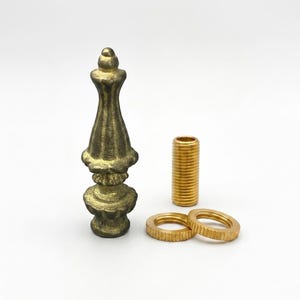 May include: A brass lamp finial with a textured, antique-style finish, accompanied by a threaded metal pipe nipple and two circular, ridged metal washers. The finial features a pointed, tiered design with a wide base and a narrow, tapered top.