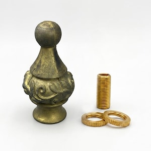 Peut inclure: Un fleuron décoratif avec une finition dorée patinée, doté d'un sommet sphérique et d'une base ornée de volutes en relief. L'ensemble comprend un mamelon fileté en laiton et deux écrous circulaires en laiton assortis pour le montage.