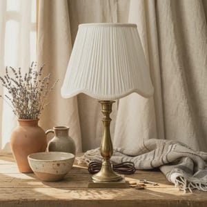 May include: A brass table lamp with a pleated white lampshade. The lamp is on a wooden table with a vase of lavender, a small bowl, and a decorative jar. A blanket and key are also on the table.