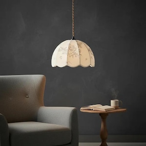 May include: Cream-colored pendant light with a floral pattern. The lampshade has a scalloped edge and hangs from a brown and white twisted cord. The light is suspended above a small wooden table with a book and a mug.