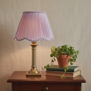 Puede incluir: Una lámpara de mesa de latón con una pantalla plisada de color lila. La lámpara está sobre una mesa de madera oscura junto a una pequeña planta en maceta y dos libros. La lámpara tiene una columna estriada y un borde festoneado.