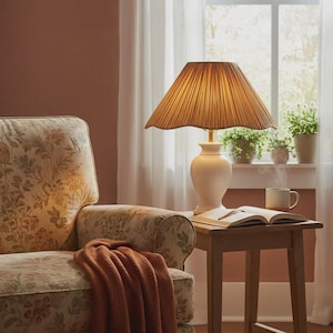 May include: A cream-colored table lamp with a pleated lampshade sits on a small wooden table. An open book and a mug are on the table. A floral patterned armchair and a rust-colored throw blanket are in the foreground. A window with plants is in the background.