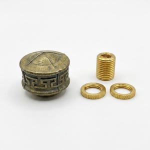 May include: A round, antique brass-colored metal knob featuring a Greek key pattern around its side and a domed top, accompanied by one threaded cylindrical metal insert and two thin, circular, serrated metal rings for assembly.