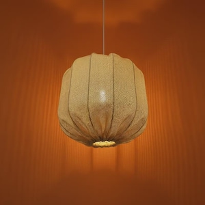 Puede incluir: Una lámpara colgante esférica beige. La pantalla está hecha de un material tejido, similar a la arpillera, con costuras verticales. La lámpara cuelga de un fino cable blanco sobre un fondo naranja. La luz está encendida, iluminando el interior.