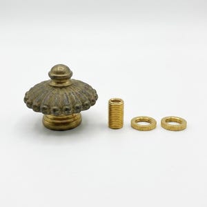 May include: A brass-toned metal furniture knob with a rounded, fluted top and a smooth base. The set includes one threaded metal cylinder and two circular, ridged metal washers, all displayed against a plain, neutral background.